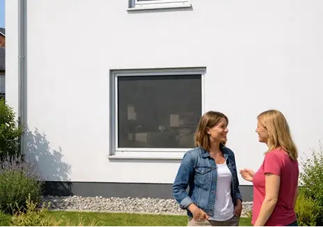 Fliegengitter am Fenster - zuverlässiger Insektenschutz fürs Zuhause Zwei Frauen stehen vor einem Hausfenster mit eingesetztem Fliegengitter und unterhalten sich