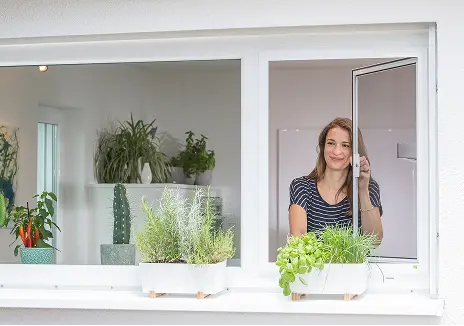 Fliegengitter-Drehfenster nach Maß für Fenster Frau öffnet ein maßgefertigtes Fliegengitter-Drehfenster an einem Fenster mit Kräutern auf der Fensterbank