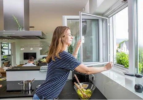 Fliegengitter-Drehfenster nach Maß für die Küche Frau öffnet ein maßgefertigtes Fliegengitter-Drehfenster am Küchenfenster