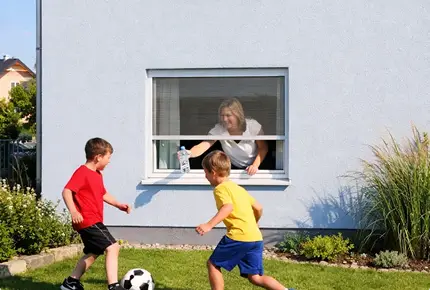 Frau bedient ein Fliegengitter-Rollo am Fenster, während Kinder im Garten Fußball spielen
