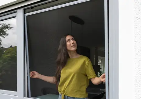 Fliegengitter-Spannrahmen für Fenster nach Maß Frau steht an einem geöffneten Fenster mit eingesetztem Fliegengitter-Spannrahmen