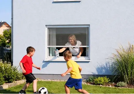Frau bedient ein Fliegengitter-Rollo am Fenster, während zwei Kinder im Garten Fußball spielen