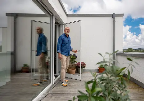 Mann öffnet eine maßgefertigte Fliegengitter-Drehtür auf einer Terrasse mit Pflanzen und Glasfront